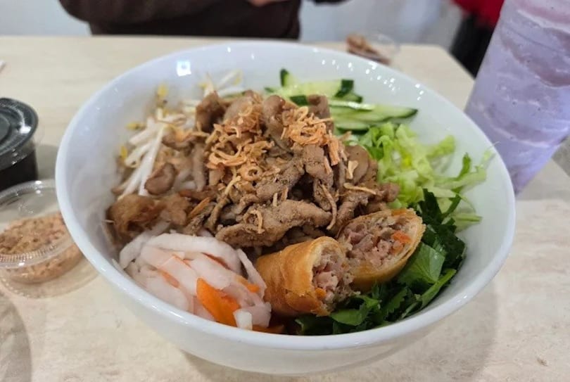 A white bowl of Vietnamese vermicelli noodles with grilled pork, spring rolls, and vegetables.
